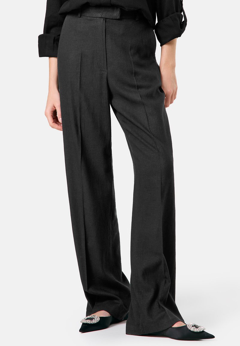 Schwarze Leinenhose mit weitem Bein, hoher Taille und sichtbaren Falten. Kombiniert mit schwarzen spitzen Schuhen, die mit einem Schmuckakzent verziert sind.