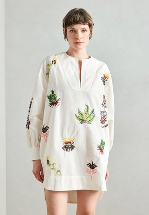 Hvid bomulds-tunika med V-udskæring og lange ærmer, prydet med farverig blomstret broderi i forskellige former og designs. Kanten er let buet.