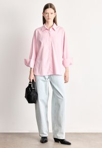 Camisa oversized às riscas cor-de-rosa e brancas com botões e mangas arregaçadas, combinada com jeans de perna larga azul-claro e uma mala preta.