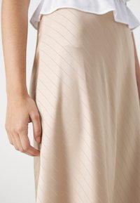 Jupe en tissu beige présentant des rayures diagonales, une texture lisse et une taille ajustée. Une main touche légèrement la surface du tissu.