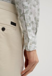 Weißes Hemd mit grünem Blumenmuster, ausgestattet mit einer geknöpften Manschette. Beigefarbene Hose mit einem schwarzen Logo-Tag und einer geknöpften Tasche.