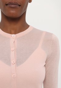 Lys rosa cardigan med en myk tekstur, med rund hals, seks knappelukking og gjennomsiktige paneler på skuldrene.