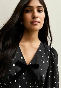 Robe noire avec des motifs d'étoiles blanches, ornée d'un accent de nœud en velours au niveau du cou. Tissu lisse avec un léger éclat, manches bouffantes longues.