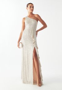 Vestido de noche de un solo hombro en color champán, silueta ajustada, detalle asimétrico de volantes, abertura alta en el muslo, combinado con un bolso de mano blanco.