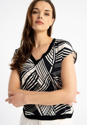 Kobieta o długich brązowych włosach, ubrana w krótki rękaw czarno-białej bluzki z geometrycznym wzorem, ze skrzyżowanymi ramionami, na jednolitym białym tle.