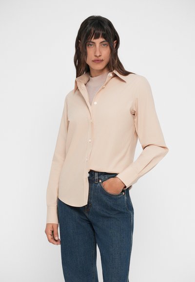 Light peach button-down shirt with a classic collar and long sleeves, made of smooth fabric. Paired with dark blue jeans featuring visible stitching.