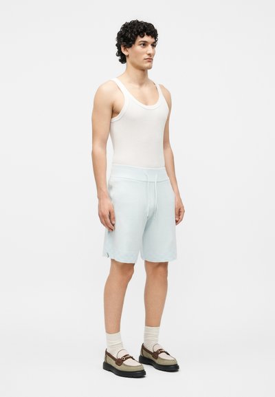 Νέος άνδρας με σγουρά μαλλιά, φοράει λευκό φανελάκι, γαλάζιες βερμούδες, μπεζ κάλτσες και καφέ μοκασίνια, στέκεται μπροστά σε απλό φόντο.