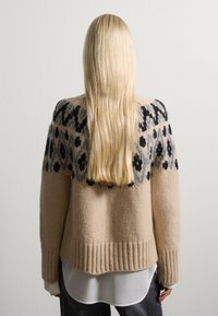 Person mit langen blonden Haaren, der einen beigen Strickpullover mit schwarz-grauem Blumenmuster, ein weißes Hemd darunter und dunkle Hosen trägt, von hinten angesehen.