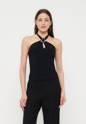 Femme portant un haut noir sans manches style dos nu avec un anneau argenté et un pantalon noir, debout devant un fond clair uni.