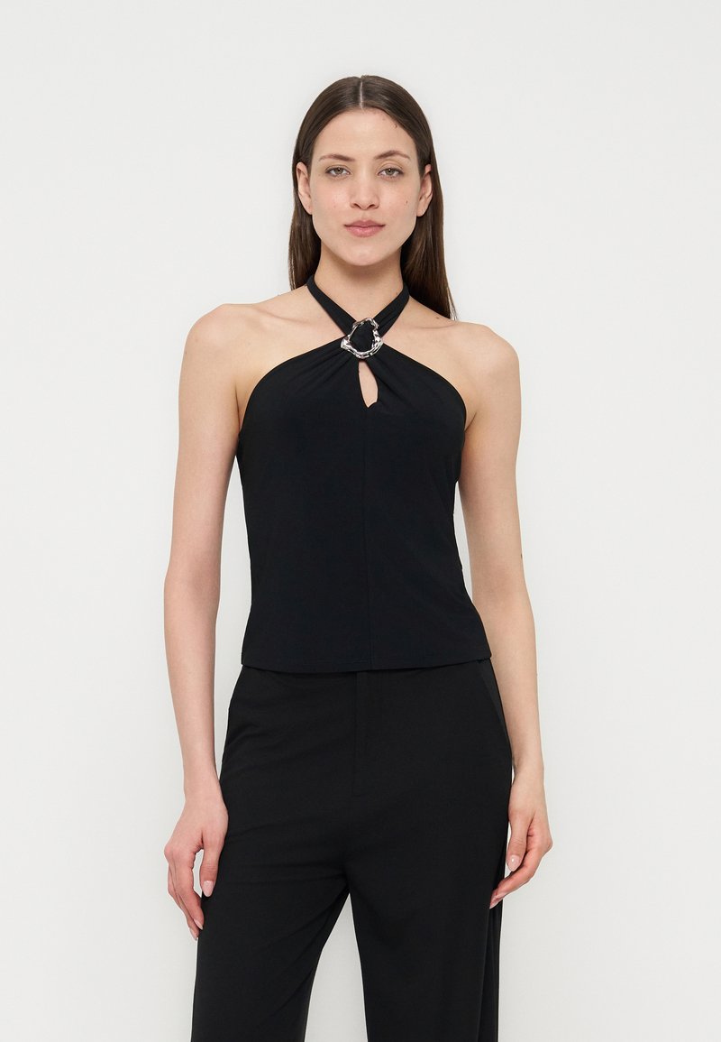 Γυναίκα που φοράει μαύρο αμάνικο τοπ με λεπτομέρεια ασημένιου δακτυλίου και μαύρο παντελόνι, όρθια μπροστά σε απλό φωτεινό φόντο.