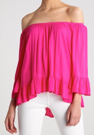 Jasnoróżowa bluzka z odkrytymi ramionami, długimi rękawami i marszczonym dołem, noszona z dopasowanymi białymi spodniami, ukazująca górną część tułowia i ręce.