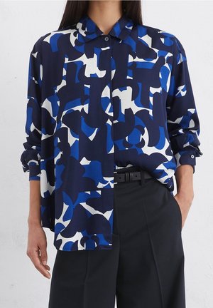 Vrouw draagt een loszittend overhemd met knopen met een abstract patroon in marineblauw, blauw en wit, gecombineerd met een zwarte, hoog getailleerde broek en riem.