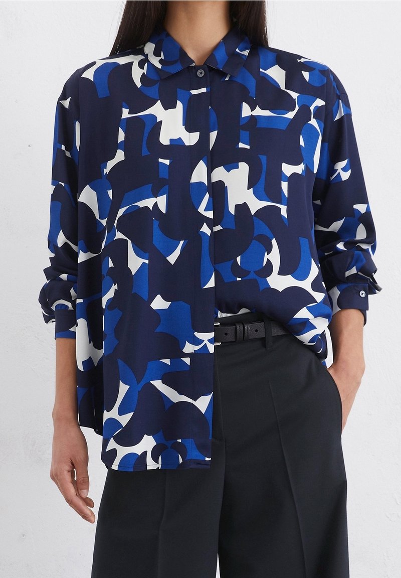Femme portant une chemise ample à boutons avec un motif abstrait bleu marine, bleu et blanc, associée à un pantalon noir taille haute et une ceinture.