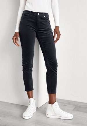 Person wearing black cropped pants, white long-sleeve top, and white high-top sneakers standing on light wood floor.