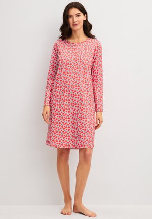 Vrouw staat blootsvoets en draagt een roze jurk met lange mouwen tot op de knie, met een rood en grijs hartjespatroon op een effen achtergrond.