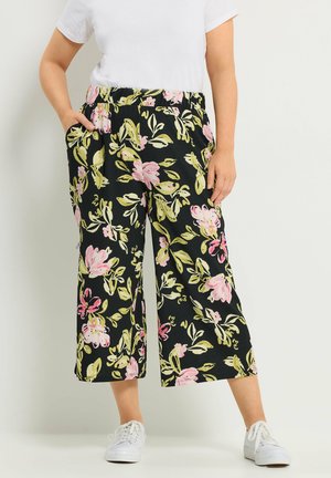 Person, die schwarze, weit geschnittene, kurz geschnittene Hose mit rosa und grünem Blumenmuster, weißes T-Shirt und weiße Turnschuhe trägt und mit den Händen in den Taschen steht.