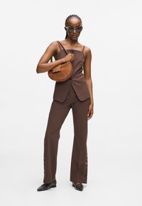 Brun skræddersyet vest og flagrende bukser med knapdetaljer, parret med en brun skuldertaske og sorte nitterede sko. Modellen bærer oversized solbriller.