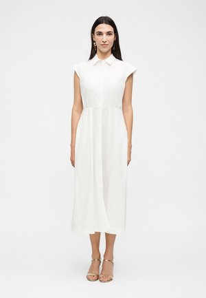 Hosszú sötét hajú nő ujjatlan fehér galléros midi ruhában és aranyszínű, nyitott orrú magassarkú cipőben, egy egyszínű fehér háttér előtt áll.