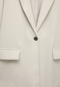 Gros plan d'un blazer beige avec un bouton noir unique et deux poches avant à rabat, montrant le revers et les détails des coutures.