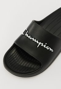 Sandalo scivoloso in gomma nera con superficie testurizzata, dotato di un logo bianco "Champion" sulla fascia e suola rigata per una miglior aderenza.