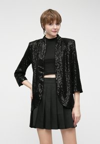 Crni sako s aplikacijama od šljokica, opuštenog kroja, skraćenim rukavima i otvorenim prednjim dijelom, u kombinaciji s crnim kratkim topom i nabranom mini suknjom.