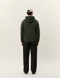 Olivovo zelená mikina s veľkou kapucňou, nosená na tmavých, voľne strihaných džínsoch. Model má na hlave svetlošedú čiapku. Jednoduchý, minimalistický dizajn.