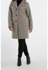 Vrouw in een knielange, grijze, gestructureerde wollen jas, dichtgeknoopt, gecombineerd met zwarte broeken en zwarte enkellaarsjes met hak.
