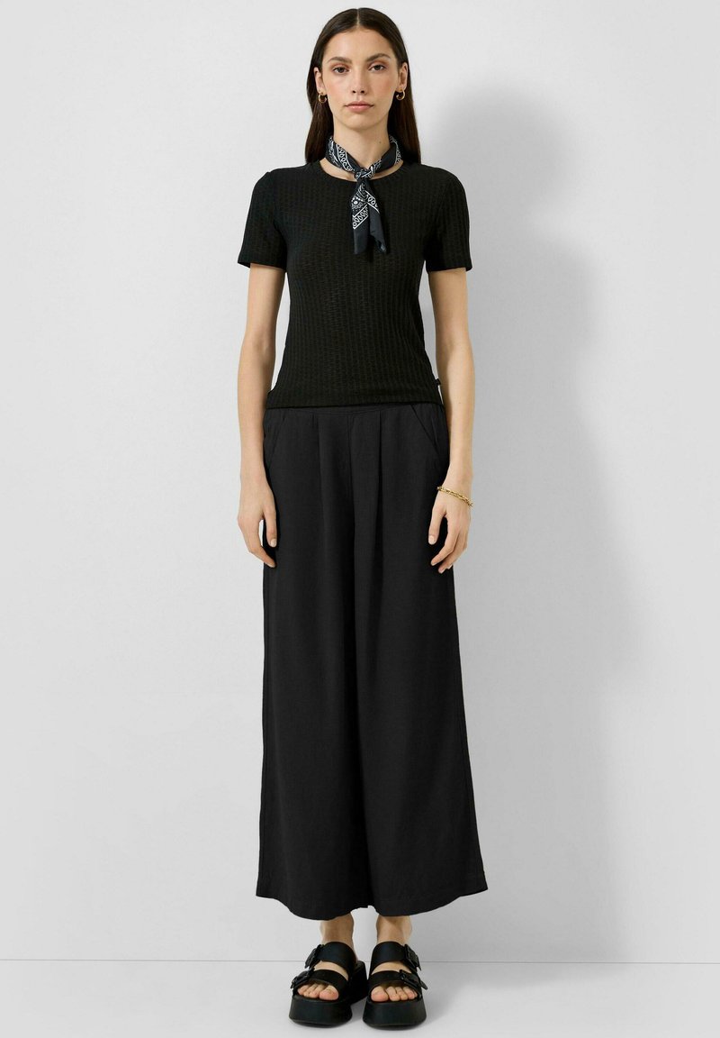 Femme debout portant un haut noir à manches courtes, un pantalon noir à jambes larges, des sandales noires et un foulard noir à motifs autour du cou.