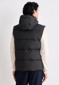 Dark puffer vest with a hood, featuring quilted sections, smooth texture, and a relaxed fit, worn over a light-colored sweatshirt.