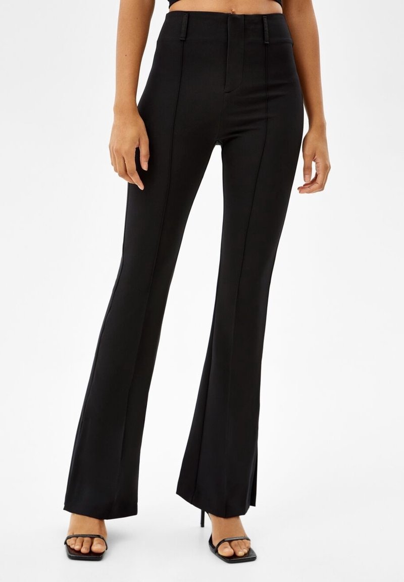 Svarta högmidjade utsvängda byxor med framseam och små sidoflikar, bärs med svarta remsandaler med fyrkantig tå och hög klack.