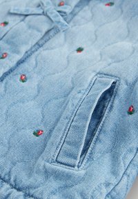 Hellblauer gesteppter Stoff mit kleinen bestickten roten und grünen Blumen und einem rechteckigen Taschen-Detail an der Kleidung.