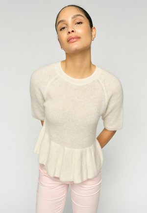 Kvinde med mørkt hår bundet tilbage iført en hvid strik-top med korte ærmer og flæsekant samt lyserøde bukser, stående foran en ensfarvet baggrund.