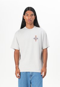 Lys grå bomull T-skjorte med korte ermer, rund hals, og et lite flerfarget grafisk design på brystet. Brukes sammen med blå jeans.