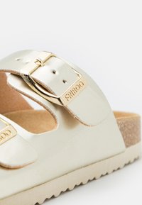Sandal s kovovými striebornými detailmi a zlatými akcentami má korkovú podrážku a nastaviteľné popruhy s prackami. Plochý dizajn s zaoblenými hranami.