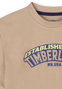 Beige katoenen t-shirt met een ronde halslijn en korte mouwen. Heeft een grote grafische tekst in blauw en geel die zegt "GESTICHTE TIMBERLAND NH. VS."