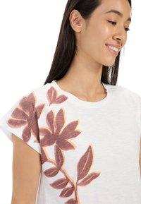 Weißes T-Shirt mit kurzen Ärmeln, das ein großes, abstraktes Blumenmuster in Bordeauxrot mit orangenen Akzenten auf der linken Seite zeigt. Weiche Textur.