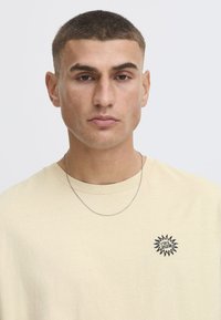 Beige bomulds t-shirt med rund halsudskæring, der har et sort broderet soldesign på venstre bryst og en sølv kædehalskæde.