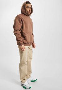 Felpa marrone con tasca frontale e cappuccio con coulisse, abbinata a pantaloni beige e sneakers bianche con dettagli verdi.