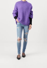 Person som bär en lila oversized tröja över svarta ärmar, trasiga ljusblå skinny jeans och svarta spetsskor med dubbla remmar.