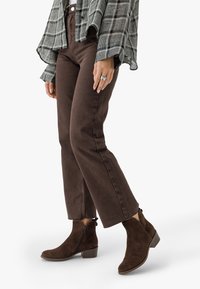 Kvinna som bär bruna korta byxor, mörkbruna mockastövlar med låg klack och en lös långärmad skjorta i rutig design.