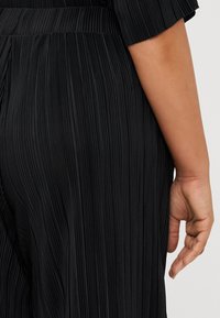 Close-up of a person's back wearing a black pleated dress, with a visible hand resting lightly on the fabric.