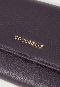 Portefeuille en cuir violet avec une surface texturée et le logo doré "Coccinelle". Design épuré avec des accents de couture visibles.
