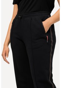 Černé tepláky vyrobené z měkké látky, s páskem na stažení, bočním zipovým kapsou a kontrastními pruhy podél nohavic.