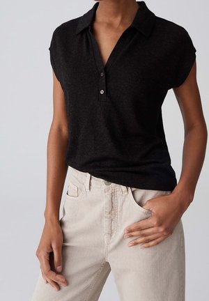 Femme portant un polo noir à manches courtes rentré dans un pantalon beige clair taille haute, une main dans la poche.