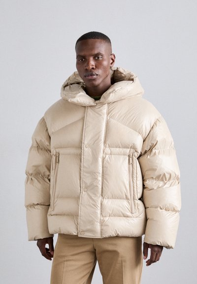 Doudoune beige avec un design matelassé, grande capuche et poches zippées. Texture lisse et brillante avec une coupe ample.