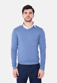 Giovane uomo in piedi di fronte, che indossa un maglione azzurro chiaro a V sopra una camicia bianca con colletto e jeans blu scuro.