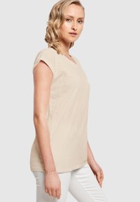 Beige t-paita lyhyillä hihoilla, jossa on V-pääntie ja huomaamaton printti. Valmistettu pehmeästä puuvillakankaasta. Yhdistetty valkoisiin housuihin.