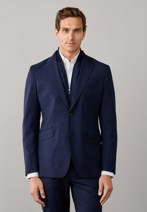 Ein Mann trägt eine marineblaue Anzugjacke mit einem marineblauen Reißverschluss-Innenlayer und einem weißen Hemd und steht vor einem einfarbigen hellgrauen Hintergrund.