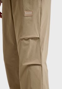 Beige bukser lavet af letvægtsstof, med et syet design og en lille, tekstureret mærke-patch på siden.