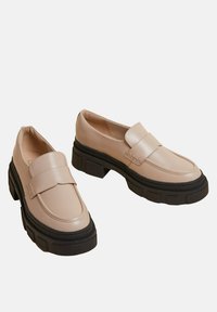Mocassins synthétiques beiges avec de épaisses semelles en caoutchouc noir, dotés d'une large sangle sur le dessus du pied et d'un design à bout rond.